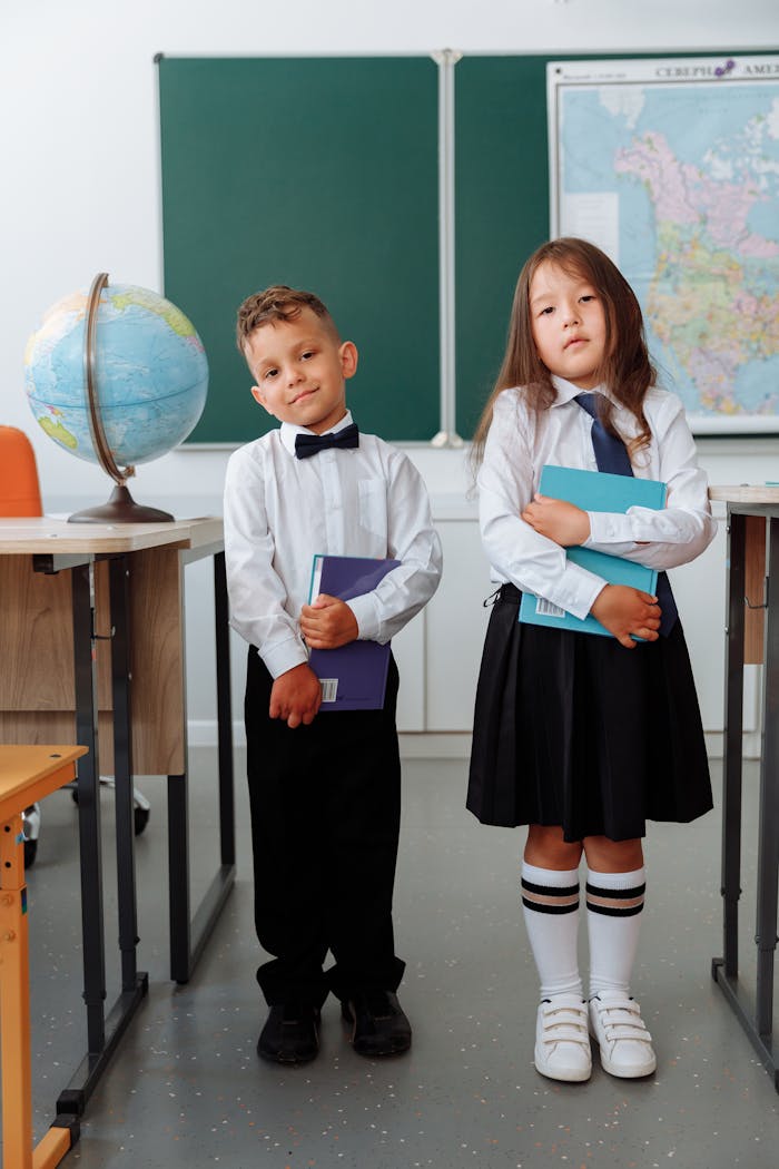 Two children in school uniforms standing and holding books in a classroom setting.